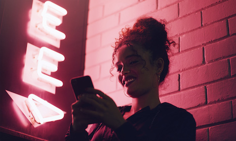 A young lady standing next to a neon sign texting on a phone